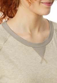 Light beige sweatshirt with a ribbed gray neckline. Features delicate stitching along the seams and a soft texture.