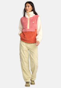 Fleece pullover in roze, crèmekleurig en oranje met hoge kraag, knoopsluiting en voorkantzak; gecombineerd met beige cargobroek.