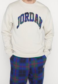 Felpa color crema con la scritta "JORDAN" in blu e motivo a quadri, abbinata a pantaloni a quadri con motivi blu, verdi e rossi.