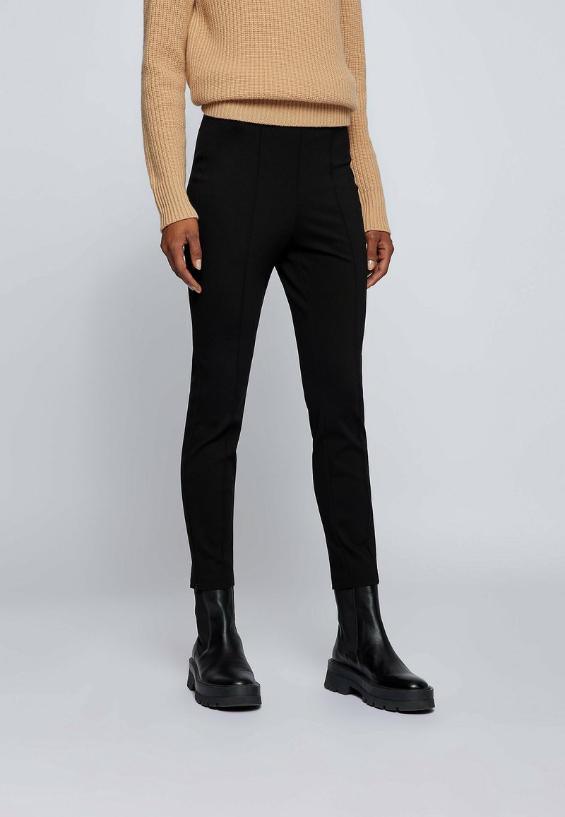Personne portant un pantalon noir ajusté, des bottines noires en cuir et un pull en tricot beige, debout devant un fond uni clair.