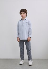 Camicia a righe azzurro chiaro a maniche lunghe con colletto e patta con bottoni; abbinata a pantaloni grigi e sneakers bianche, in piedi su un pavimento grigio.