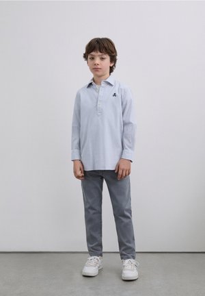 Camisa de manga larga a rayas azul claro con cuello y tapeta de botones; combinada con pantalones grises y zapatillas blancas, de pie sobre un suelo gris.