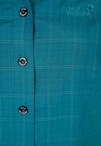 Tyrkysová košile na knoflíky s kostkovaným vzorem, s černými knoflíky s vyraženým logem. Měkká, strukturovaná látka s jemným leskem.