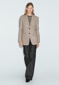 Blazer beige su misura con due tasche frontali e bottoni neri, indossato sopra una camicia a righe bianco e nera, abbinato a pantaloni larghi scuri.