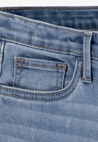 Blugi din denim de culoare albastru deschis, cu o textură ușor estompată. Au un buzunar mic și cusături contrastante de-a lungul taliei și marginilor buzunarului.