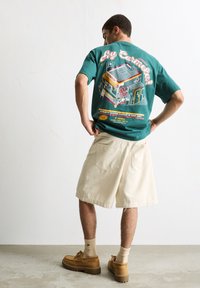 T-shirt en coton teal avec un design graphique au dos, associé à un short beige et des chaussures marron. Manches courtes, coupe décontractée.
