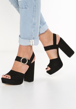 Stopy w jasnoniebieskich podwiniętych dżinsach oraz czarnych sandałach na wysokim obcasie z dużą okrągłą klamrą na pasku wokół kostki.