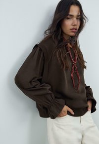 Maglione marrone con maniche voluminose, polsini arricciati e scollo texturizzato. Abbinato a pantaloni bianco sporco e una sciarpa rossa con motivo.