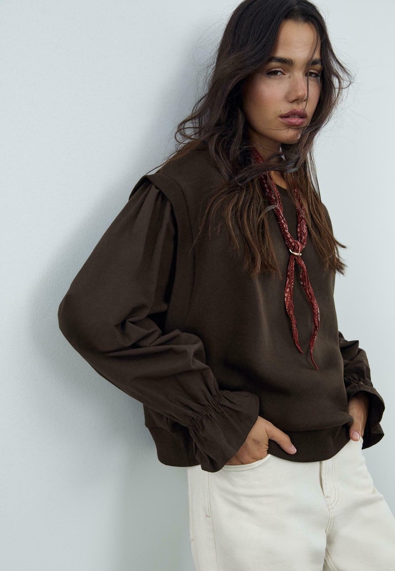 Maglione marrone con maniche voluminose, polsini arricciati e scollo texturizzato. Abbinato a pantaloni bianco sporco e una sciarpa rossa con motivo.