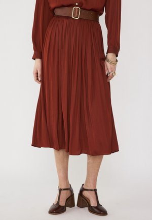 Femme portant une jupe midi plissée couleur rouille, un chemisier assorti à manches longues, une ceinture marron épaisse, des bracelets en or, des bagues et des chaussures à talons marron à bride en T.