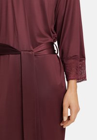 Burgundy satinrock med knytmidja, med en delikat spetskant på ärmarna och en slät, semiglansig yta.