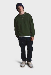 Sudadera verde de manga larga, gorro azul oscuro, vaqueros de mezclilla oscuros y zapatillas crema. La sudadera presenta un pequeño logo en el pecho.