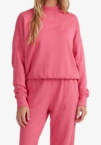 Donna che indossa un maglione a manica lunga rosa e pantaloni abbinati con "ROXY" ricamato sul petto, in piedi su uno sfondo bianco.