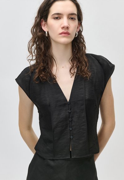 Νεαρή γυναίκα με σγουρά μαλλιά μήκους μέχρι τους ώμους, φοράει μαύρο κοντομάνικο μπλουζάκι με κουμπιά και ασημένια σκουλαρίκια κρίκους, σε απλό φόντο.