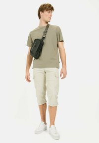 Sort crossover taske med lynlåse, grøn t-shirt, beige cargo-shorts og hvide sneakers. Modellen står sideværts på en hvid baggrund.