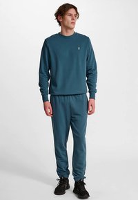 Türkisfarbener Pullover und passende Jogginghose aus weichem Stoff. Der Pullover trägt ein kleines Logo auf der Brust. Die Hose hat elastische Bündchen.
