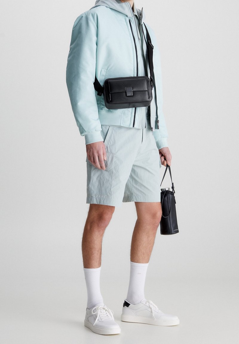 Světle modrá bomber bunda se šedou kapucí, černá krosová taška, světlé kraťasy, bílé tenisky a černá válcová taška. Čistý design, vrstvený vzhled.