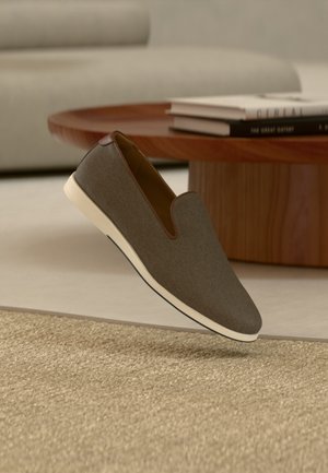 Brauner Slip-On-Freizeitschuh mit weißer Sohle, der über einem strukturierten beigen Teppich schwebt, im Hintergrund ein runder Holztisch und ein Stapel Bücher.