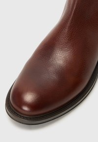 Bruine leren laars met een gladde textuur, ronde neus en subtiele glans. Beschikt over een smalle zwarte rubberen zool met stikseldetails.