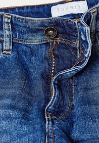 Blå denimjeans med kontrasterande orange sömnad, metallknäppning och dragkedja. Etiketten säger "ESPRIT." Tyget har en klassisk struktur.