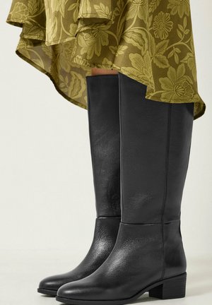 Bottes en cuir noires jusqu'aux genoux portées avec une robe verte à motif floral, présentées de mi-mollet aux pieds sur un fond clair.