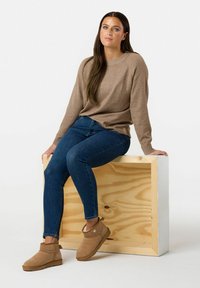 Maglione lavorato a maglia beige, jeans blu attillati e stivaletti in camoscio marrone al polpaccio. Modella seduta su un cubo di legno chiaro con una finitura naturale liscia.