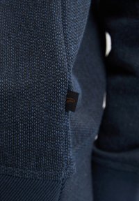 Tessuto blu navy con una trama fitta. Presenta un piccolo etichetta nera con cuciture arancioni. Vista ravvicinata che enfatizza i dettagli del materiale.