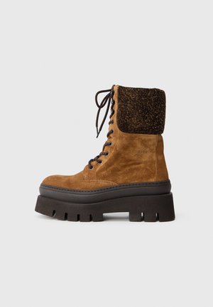 Bota de tornozelo em suede castanho com colarinho de pêlo sintético preto, frente com atacadores e sola plataforma preta robusta com detalhes recortados.