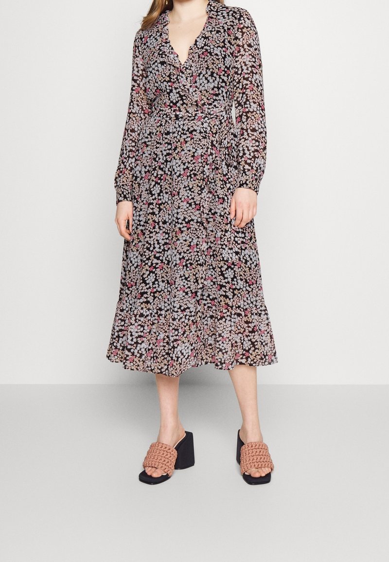Robe midi florale avec un fond noir, ornée de motifs floraux roses et blancs, manches longues et décolleté en V. Associée à des sandales à talons épais.