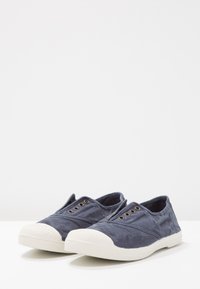 Plavetne platnene natikače z belim gumijastim zaščitnim delom za prste. Imajo tri kovinske zračne oči na vsaki strani za zračenje.