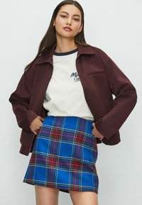 Bordeauxfarbener Reißverschlussjacke, cremefarbenes T-Shirt mit marineblauen Akzenten und blauer Karorock mit roten und gelben Details. Glatte Texturen und lässiges Design.