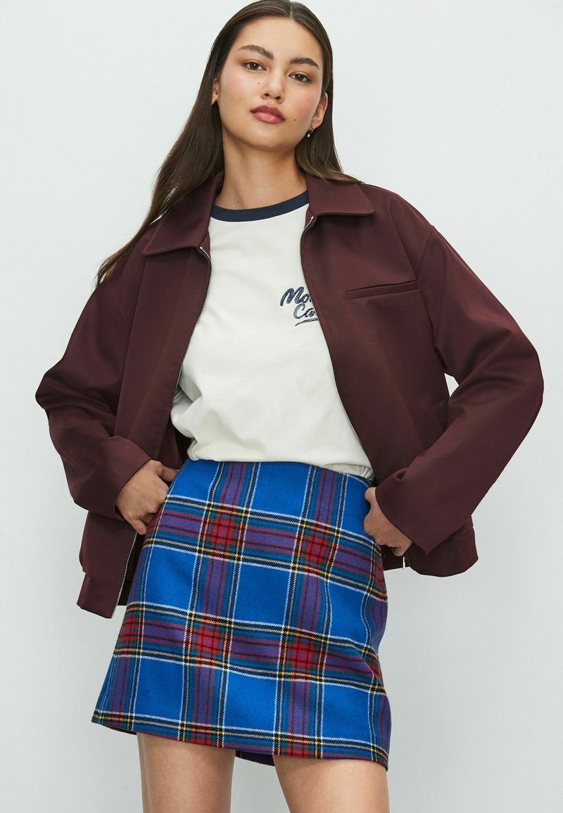 Bordeauxfarbener Reißverschlussjacke, cremefarbenes T-Shirt mit marineblauen Akzenten und blauer Karorock mit roten und gelben Details. Glatte Texturen und lässiges Design.