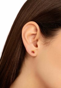 Guldpläterade örhängen med en central röd sten, omgiven av mindre klara stenar, visad på en modells örsnibb med mörkt hår.