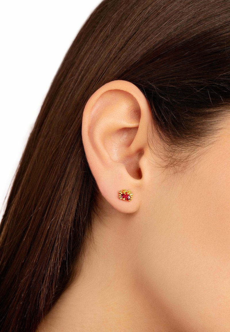 Guldpläterade örhängen med en central röd sten, omgiven av mindre klara stenar, visad på en modells örsnibb med mörkt hår.