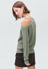 Grüner gestrickter Pullover mit schulterfreiem Design, gerippten Bündchen und unterem Saum. Getragen mit einem schwarzen Rock mit Taschen und dekorativen Nieten.