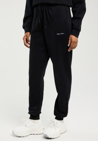 Osoba stojící proti jednoduchému světlému pozadí, oblečená v černých teplákových kalhotách se šňůrkou v pase a bílých teniskách.