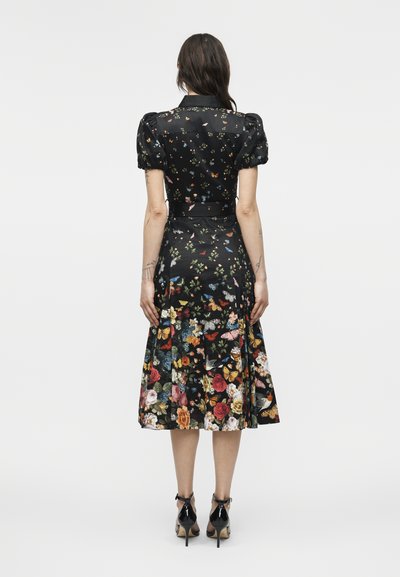 Femme portant une robe noire mi-longue avec des manches bouffantes, un imprimé floral et papillons, ainsi que des chaussures à talons noirs, debout dos au spectateur.