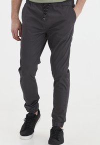 Pantalones jogger de color gris oscuro, hechos de una tela ligera, con una cintura elástica y cordón, y puños acanalados. Llevados con zapatillas negras.