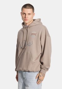 Jasnobrązowa bluza z kapturem z kieszenią kangurką, z graficznymi detalami w czerwonym i białym kolorze. Miękka, casualowa tkanina z luźnym krojem i sznurkiem w kapturze.