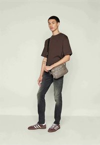 Giovane in piedi con una borsa a tracolla, indossando una maglietta marrone, jeans neri sbiaditi e sneaker bordeaux con strisce bianche.