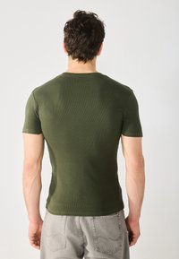 Camiseta ajustada de manga corta verde con cuello redondo, hecha de tela de canalé. Combinada con pantalones grises y ligeros.