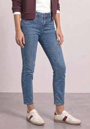 Personne portant un jean en denim bleu, des baskets blanches avec des rayures bordeaux, une chemise blanche et une veste bordeaux debout sur un sol gris.