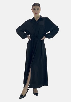 Abito lungo nero a maniche lunghe con abbottonatura frontale, vita cin cinata e spacco laterale, realizzato in un tessuto liscio. Indossato con tacchi a punta neri.