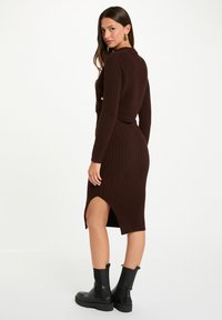 Abito knit a coste marrone con collo alto, silhouette aderente, spacco laterale e maniche lunghe, abbinato a stivali neri alla caviglia.
