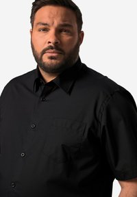 Camisa negra de manga corta hecha de una tela suave, con cuello puntiagudo y un único bolsillo en el pecho con cierre de botón.