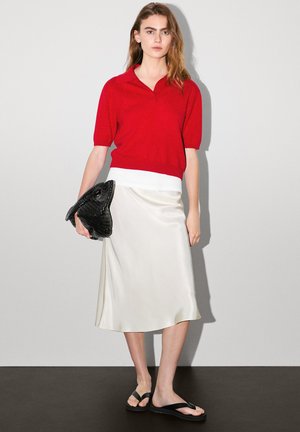 Rode polo met korte mouwen, witte tailleband, satijnen ivoorkleurige rok, zwarte getextureerde clutch in de vorm van een kikker en zwarte slippers.