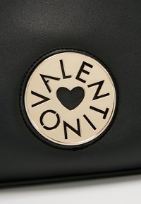Musta nahkainen käsilaukku, jossa on pyöreä logo, jossa lukee "VALENTINO" kullanvärisellä metallilla, ja keskellä sydänyksityiskohta.
