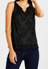 Top senza maniche nero con texture in pizzo, scollatura a chiave e dettagli in metallo dorato. Abbinato a jeans scuri per il contrasto.