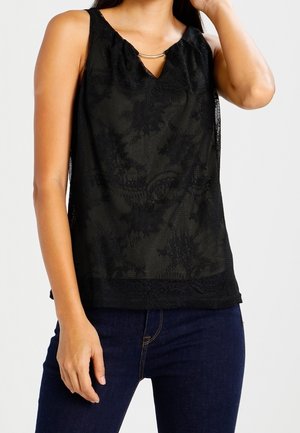 Top senza maniche nero con texture in pizzo, scollatura a chiave e dettagli in metallo dorato. Abbinato a jeans scuri per il contrasto.