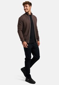 Braune Zip-Jacke mit Fischgratmuster, kombiniert mit einem schwarzen Hemd und dunkler Hose, ergänzt durch schwarze Sneaker.